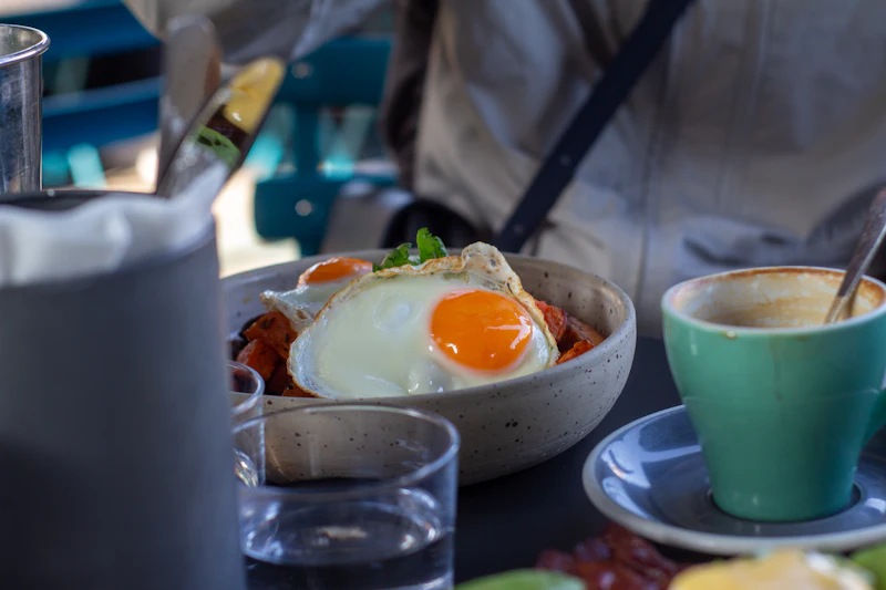 A speckled bowl of roast vegetables topped with two fried eggs, next to a green cup of coffee.