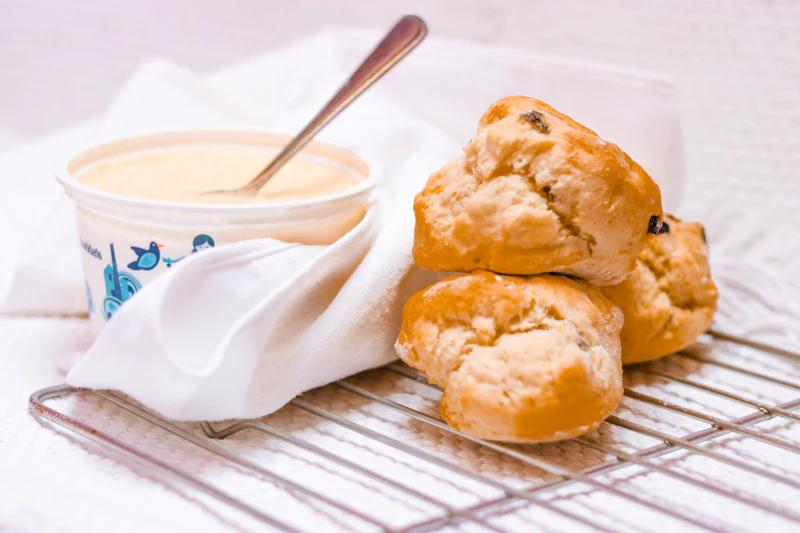 Three golden fruit scones stacked on a cooling rack beside a pot of cream and a folded white linen cloth.