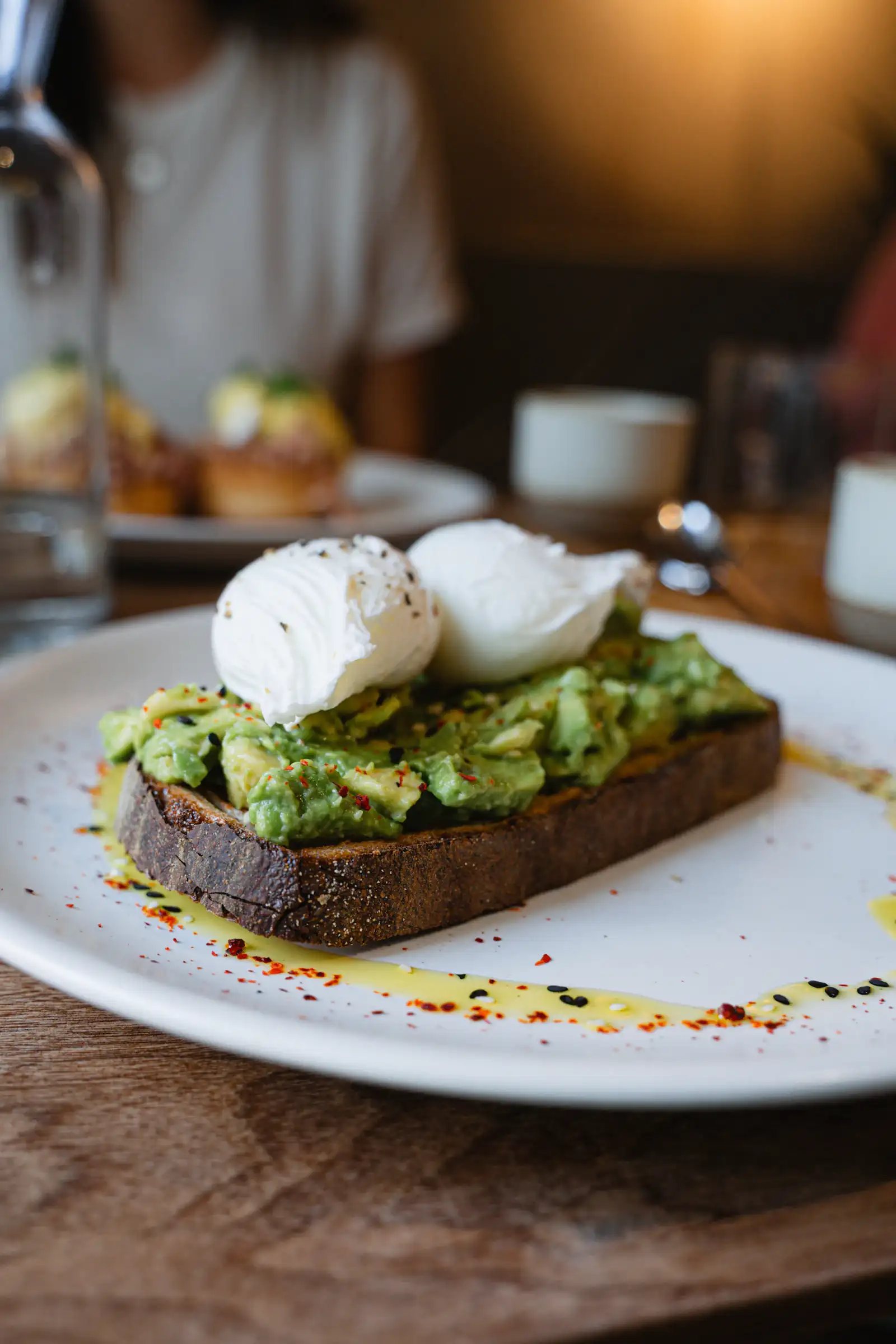 Two soft poached eggs on crushed avocado and thick toasted sourdough, finished with chilli flakes and olive oil on a white plate.