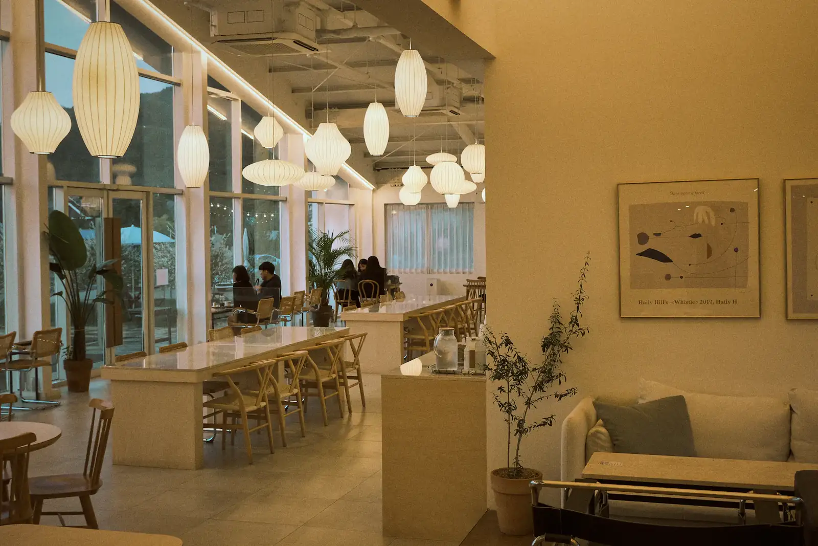 A long, glass-walled dining room glowing under paper lantern pendants, with oak tables, rush-seat chairs and a few diners by the windows.