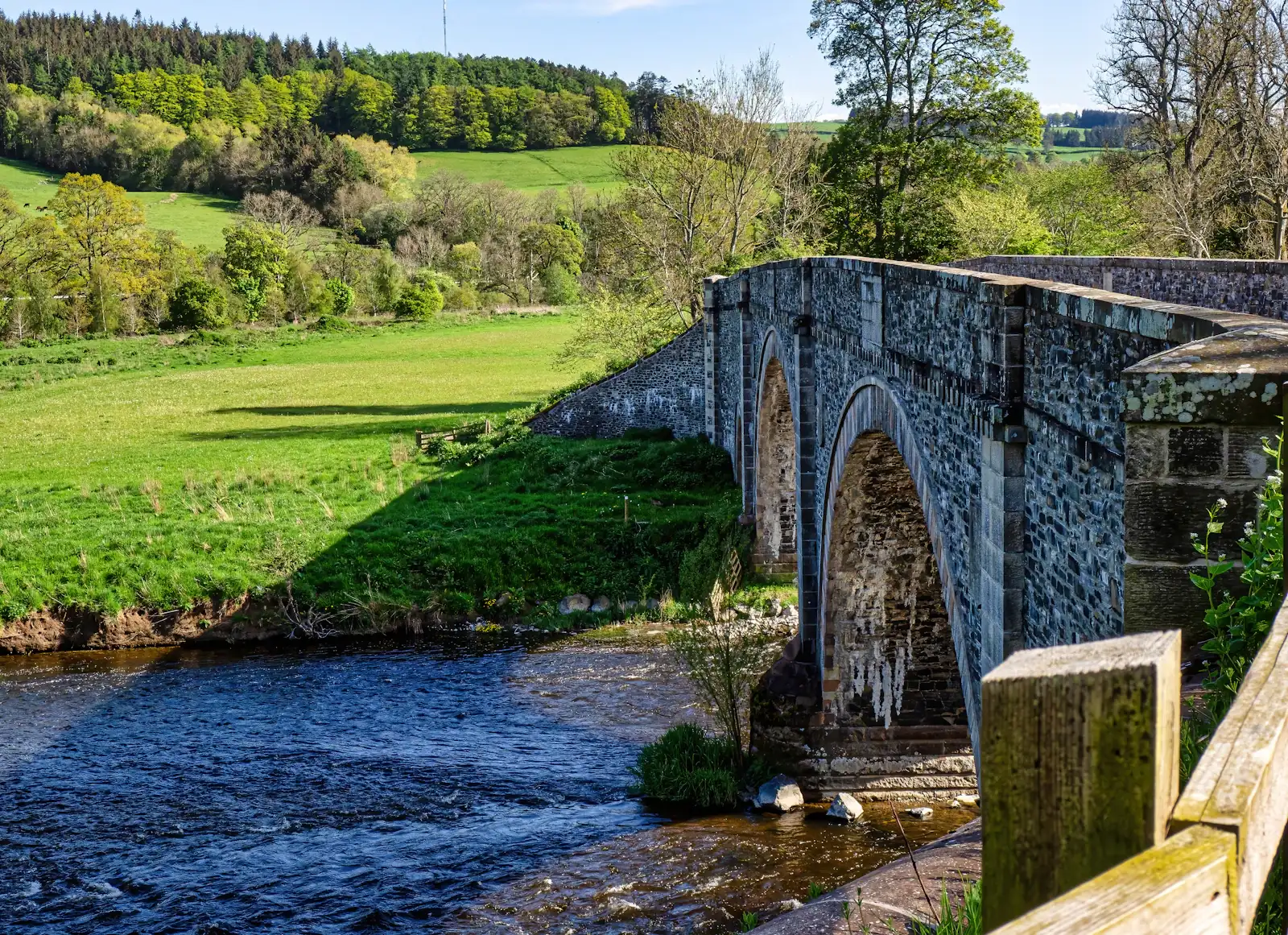 An old arched stone bridge crossing a clear, fast-flowing river between bright green fields and spring woodland.