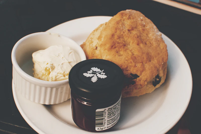 A single fruit scone on a white plate with a ramekin of clotted cream and a small jar of strawberry jam.