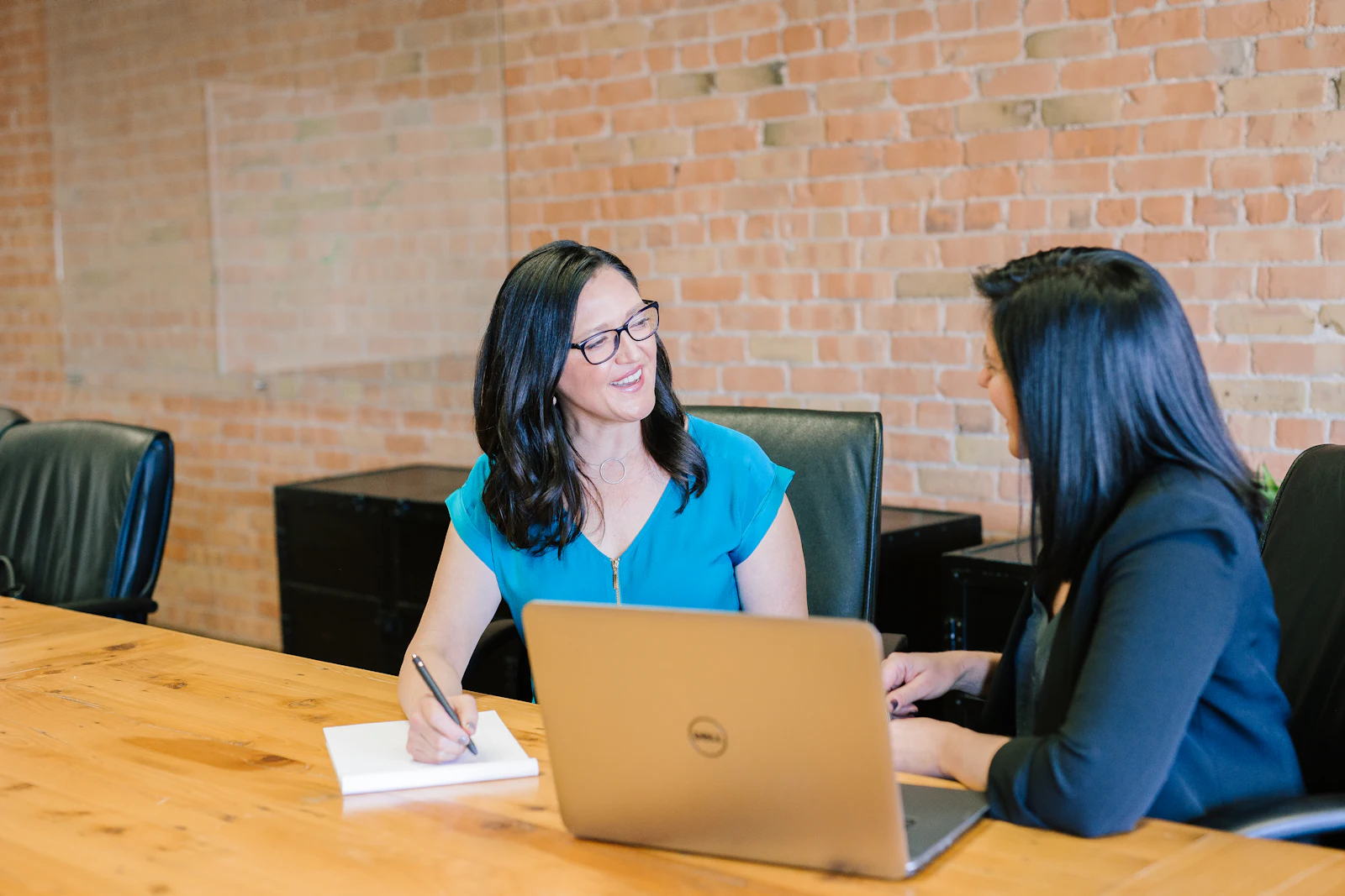 An accountant in glasses smiles and takes notes while talking through figures with a client across a wooden table, laptop open between them, in an office with an exposed brick wall.