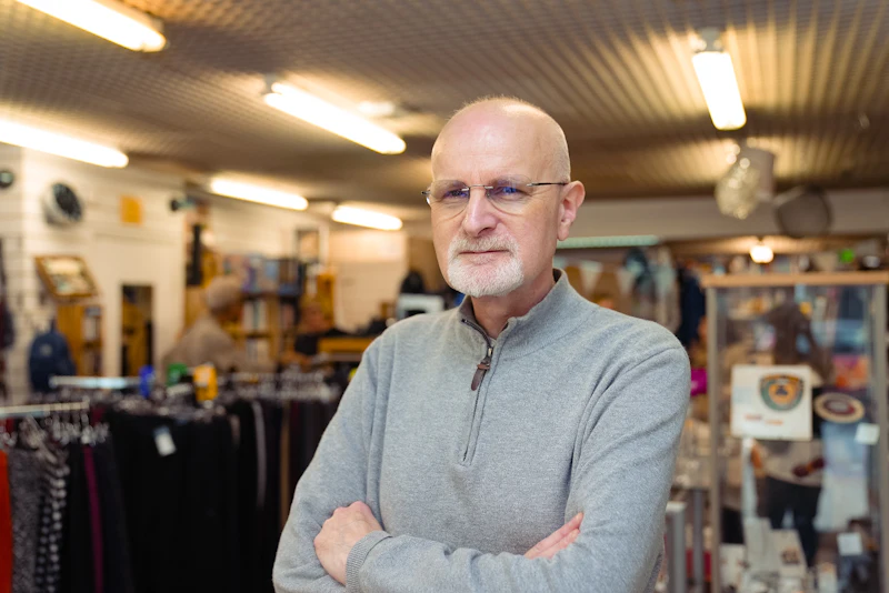 A shop owner in a grey quarter-zip jumper stands with arms folded in his clothing shop, rails of garments behind him.