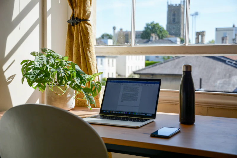 A laptop on a desk by a sunny sash window, looking out over rooftops towards a stone church tower.