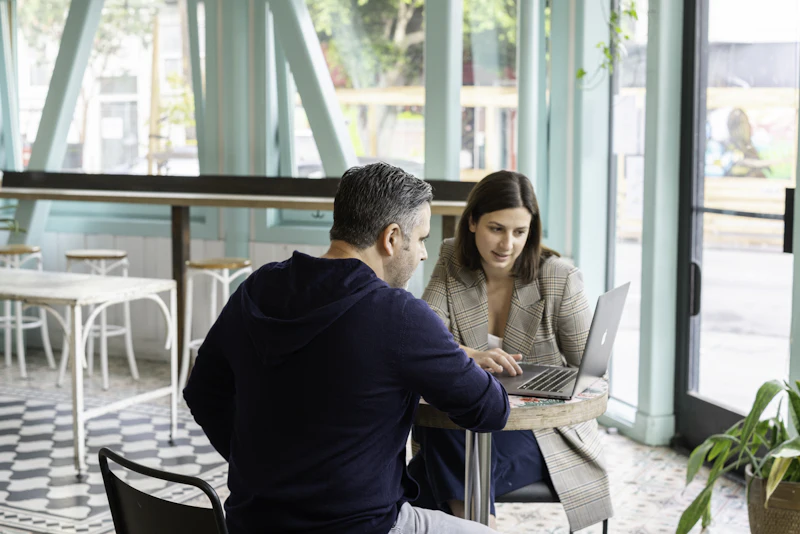 A café owner and an adviser sit at a small round table by the window, going through figures on a laptop.