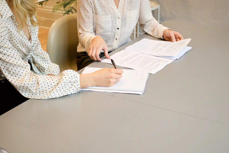 Two colleagues at a grey desk: one signs a notebook page while the other turns over a set of printed accounts.