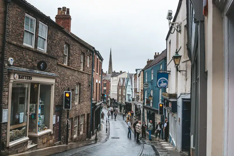 A narrow high street of stone and brick buildings on a grey day, independent shops at street level and a church spire at the far end.