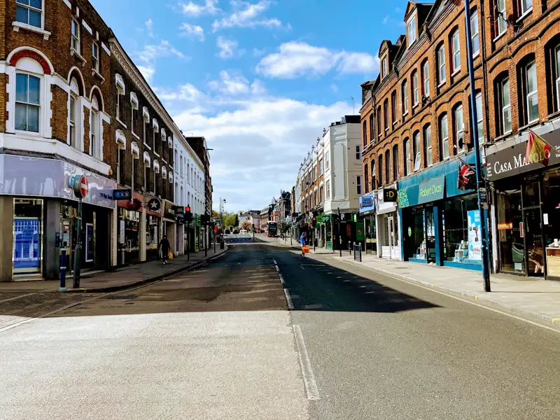 A quiet town-centre high street of brick terraces with shopfronts under a bright, cloud-flecked sky.