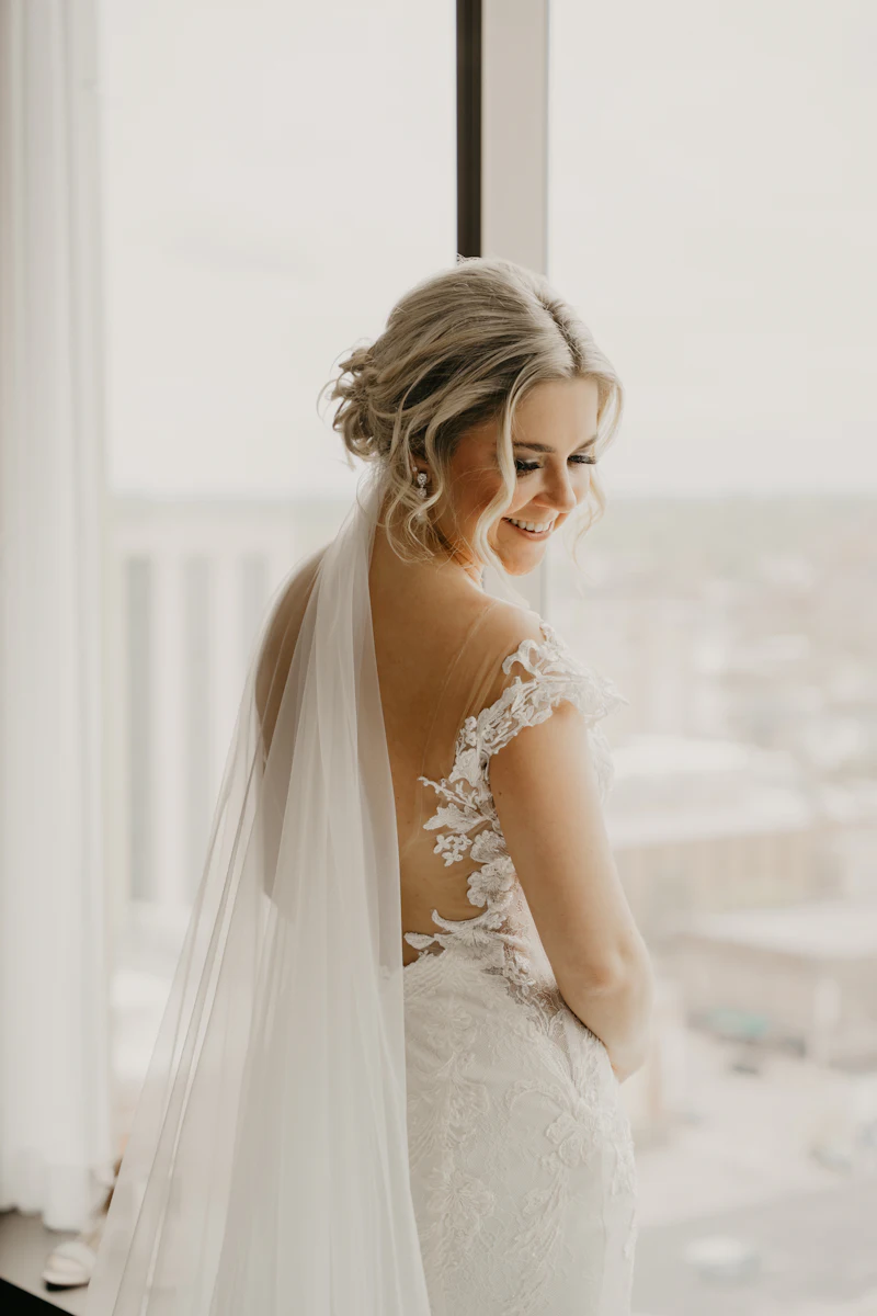 A smiling bride in a lace-backed gown and long veil looks over her shoulder by a tall window, her hair in a soft, loose updo.