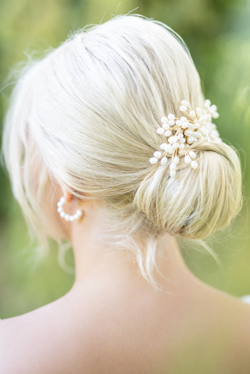 The back of a sleek platinum bridal bun finished with a delicate gold-and-pearl comb, against soft green foliage.