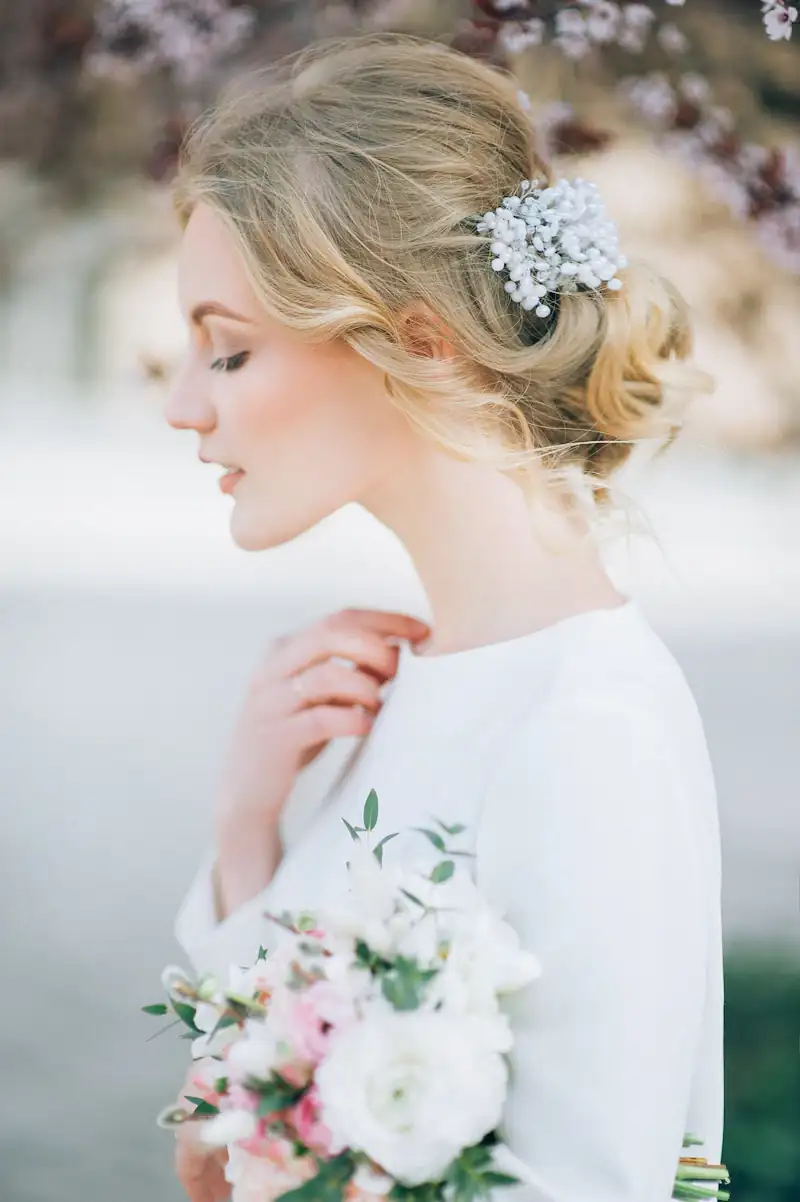 Side profile of a bride with a romantic low chignon, dressed with a cluster of pearl hairpins, holding a blush and white bouquet.