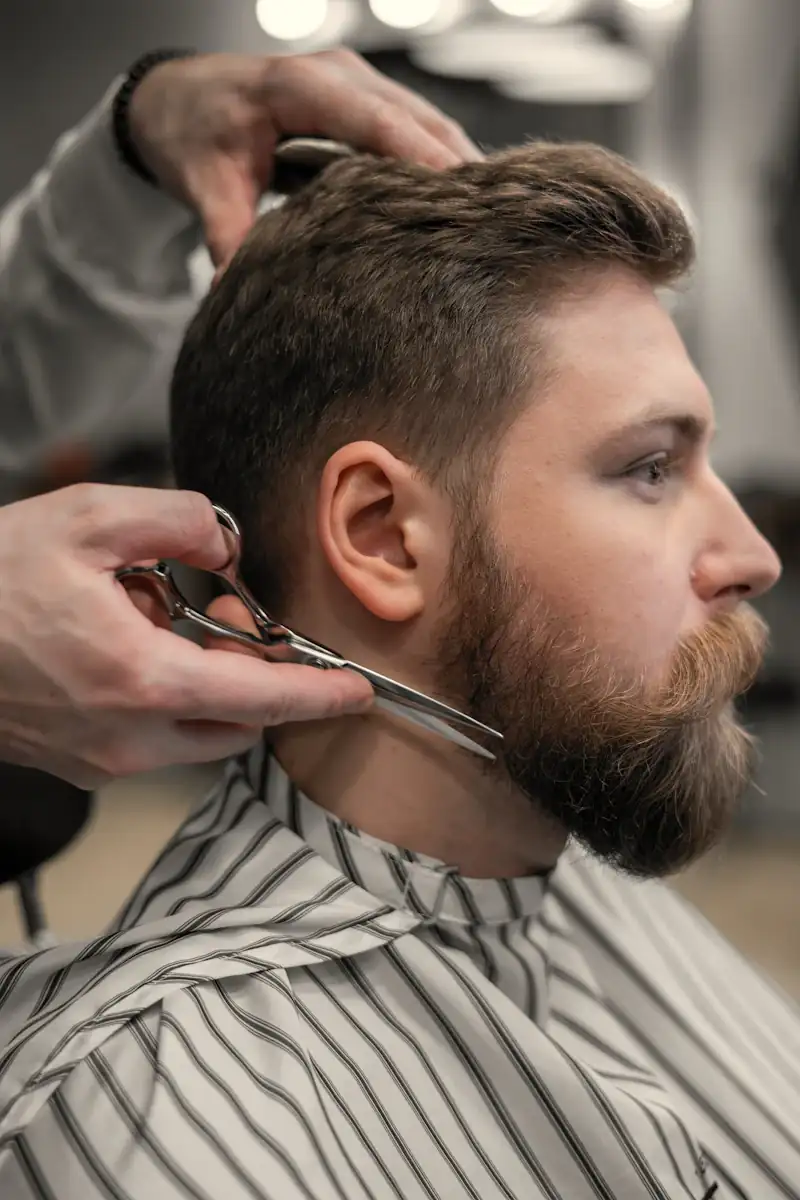 Close-up of a stylist trimming the neckline of a bearded man's short, textured haircut with silver scissors.