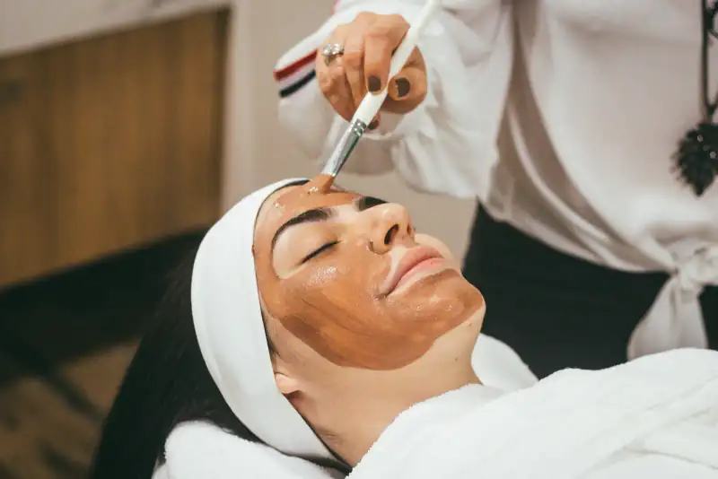 A therapist brushes a warm clay mask across a client's face as she relaxes, eyes closed, in a white headband and robe.