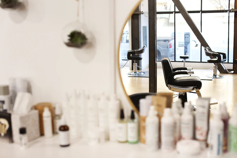 A round brass-framed mirror reflects an empty styling chair and tall windows, with organic skincare bottles lined up in front.