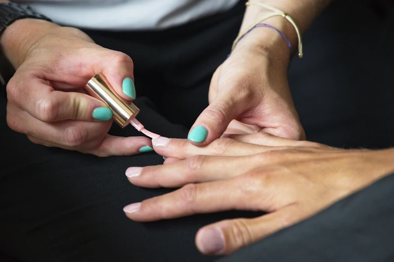 A nail technician paints a soft blush polish onto a client's nails, holding a gold-capped bottle.