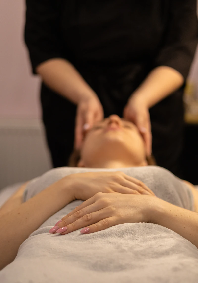 A therapist cradles a client's face during a relaxing facial massage, the client resting under a soft grey towel.