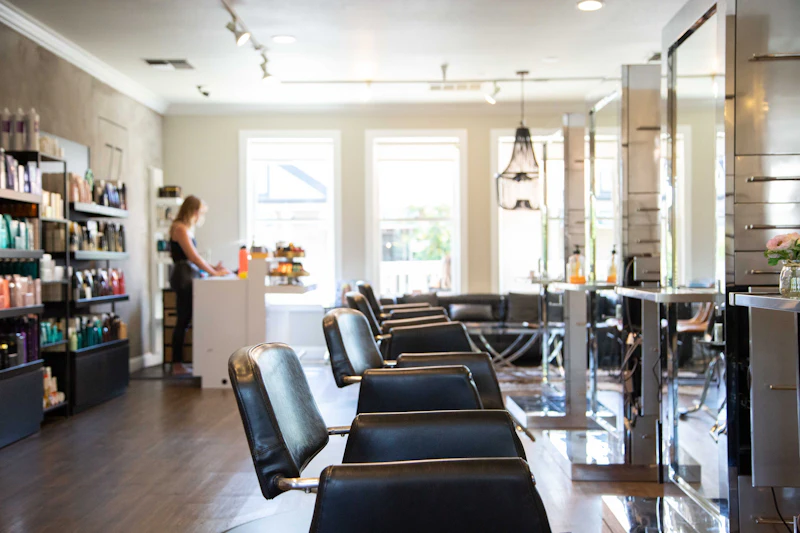 A row of black leather styling chairs facing tall mirrors, with shelves of hair-care products by the window.