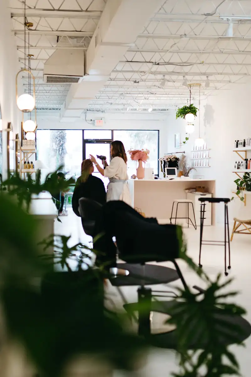 A bright, airy salon with white walls, globe pendant lights and trailing plants, a stylist at work in the background.