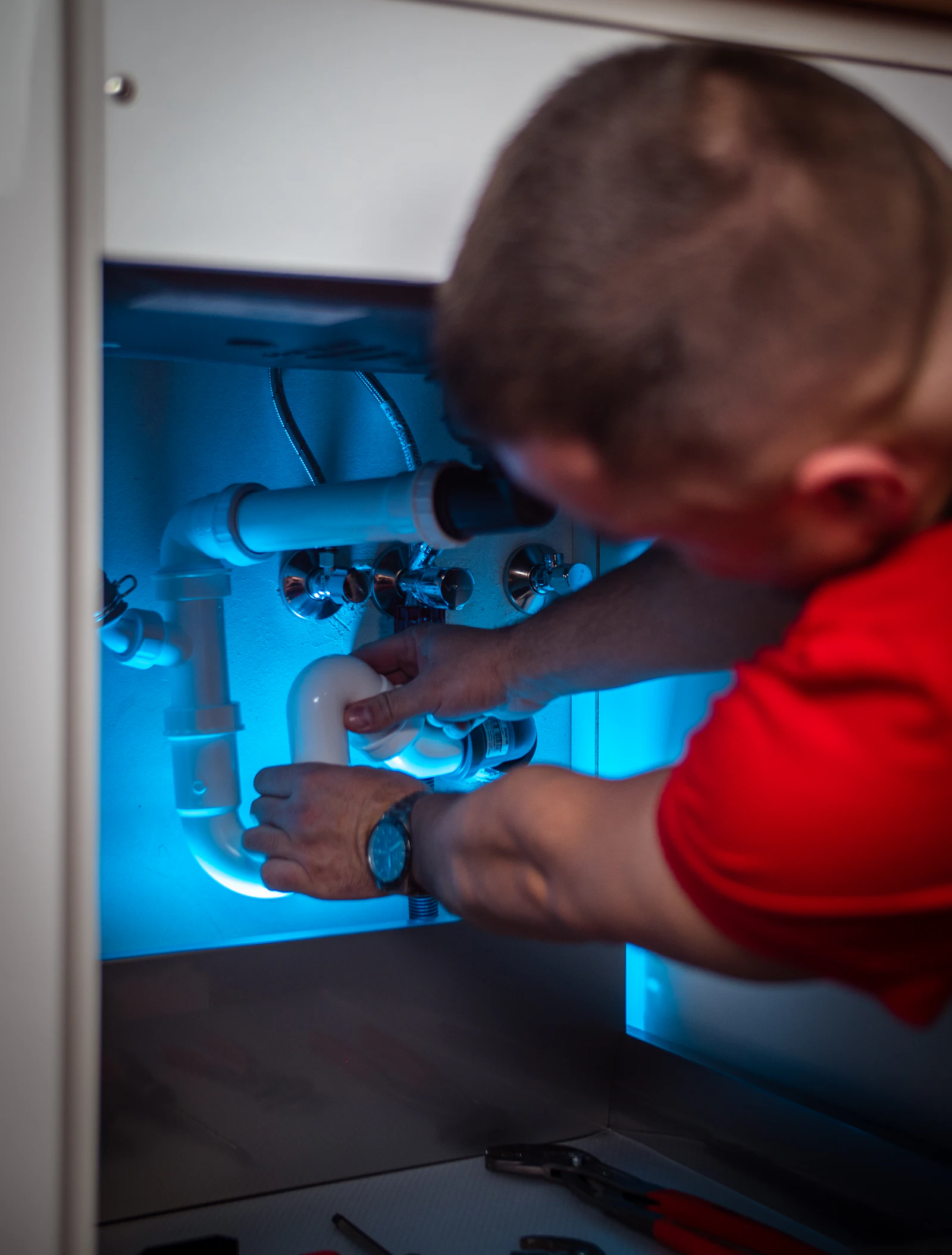 A plumber in a red shirt refits the waste pipework beneath a sink unit