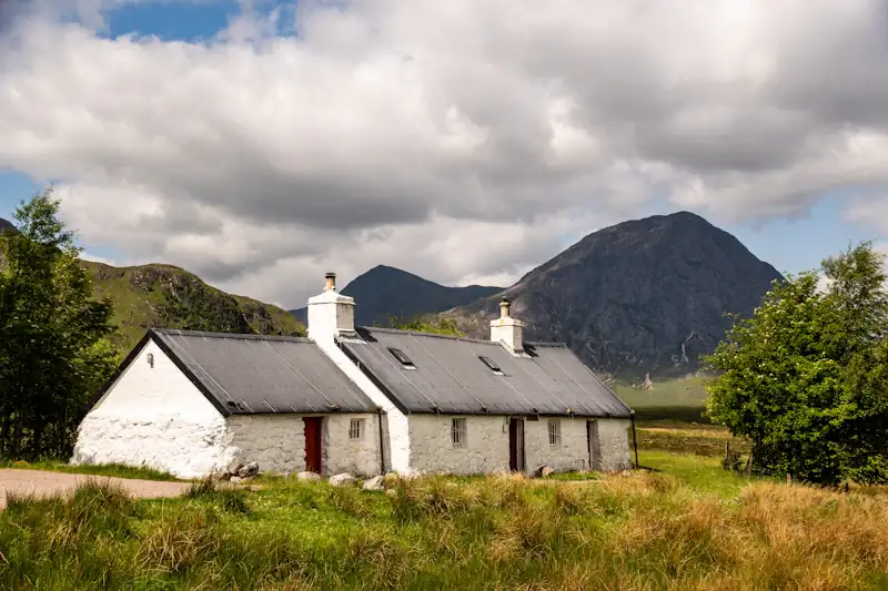 A whitewashed stone cottage with a slate roof and red door beneath hills
